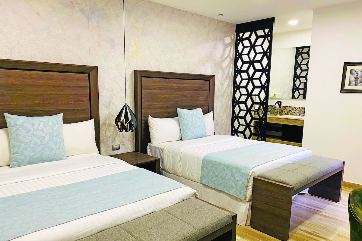 A hotel room with two beds, a wooden headboard, a nightstand, a bench, a decorative partition, and a mirror on the wall.