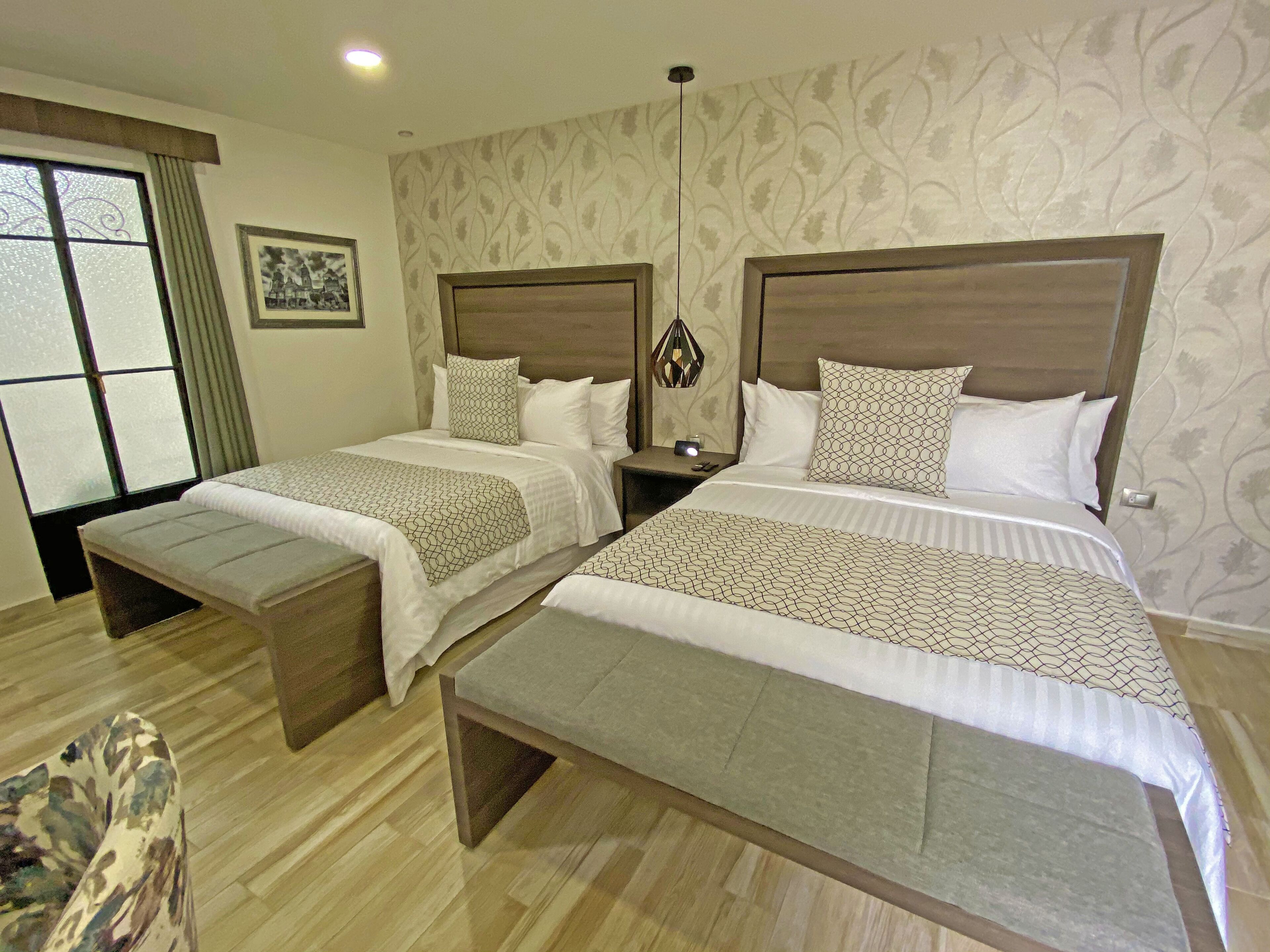 A hotel room with two beds, a bench, a nightstand, a wall-mounted lamp, and a window with curtains.