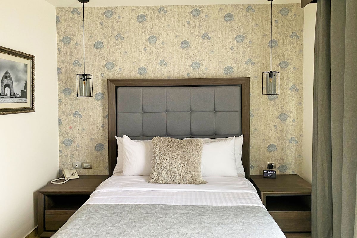 A neatly arranged bedroom with a large bed, two bedside tables, a framed picture on the wall, and a patterned wallpaper.
