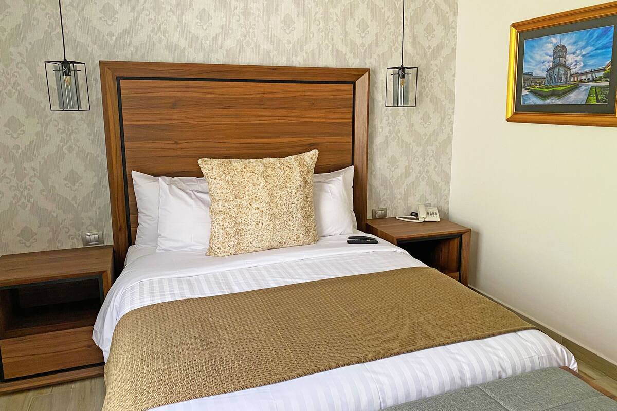 A hotel room with a wooden headboard, a bed with white and patterned bedding, a brown throw blanket, a gray pillow, and a nightstand with a phone and remote controls.