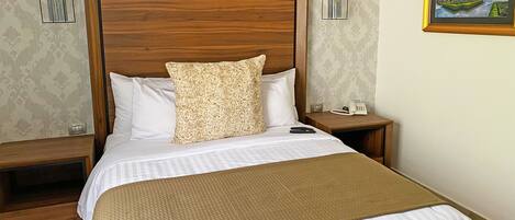 A hotel room with a wooden headboard, a bed with white and patterned bedding, a brown throw blanket, a gray pillow, and a nightstand with a phone and remote controls.