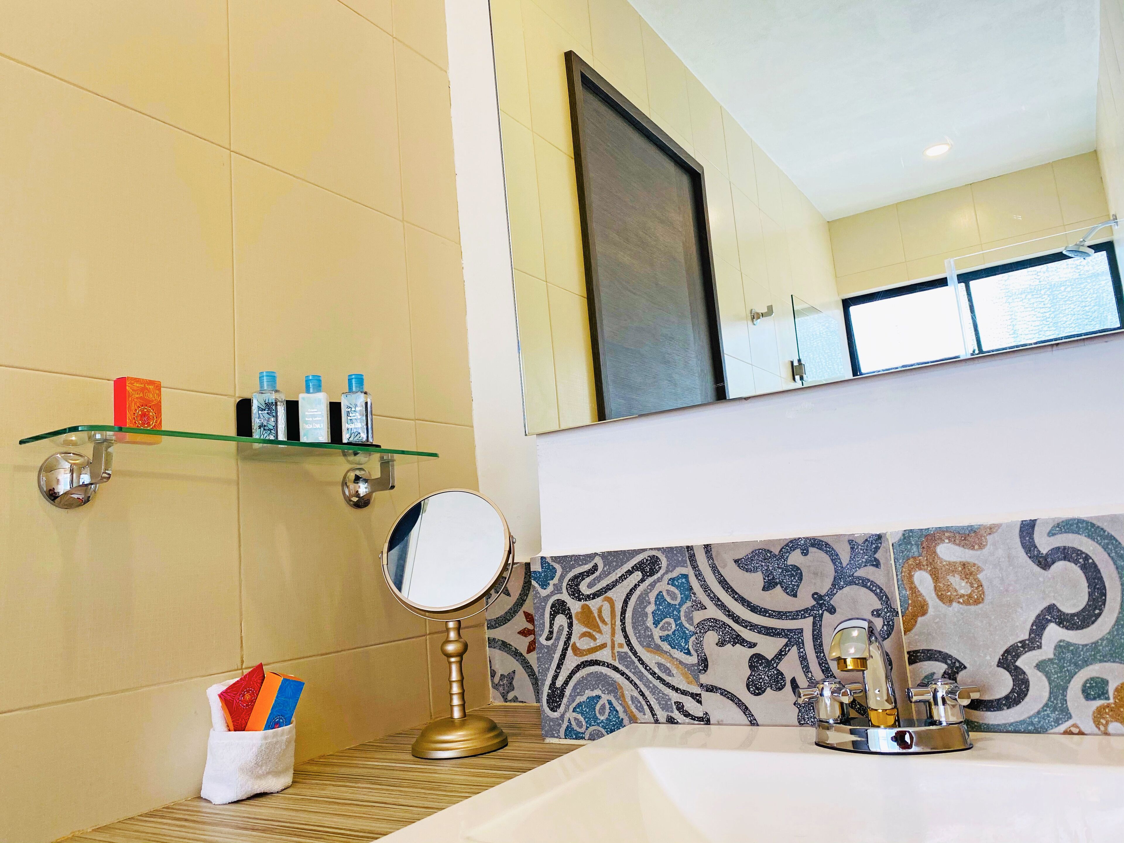 A bathroom with a tiled wall, a glass shelf with toiletries, a round mirror, and a patterned tile backsplash.