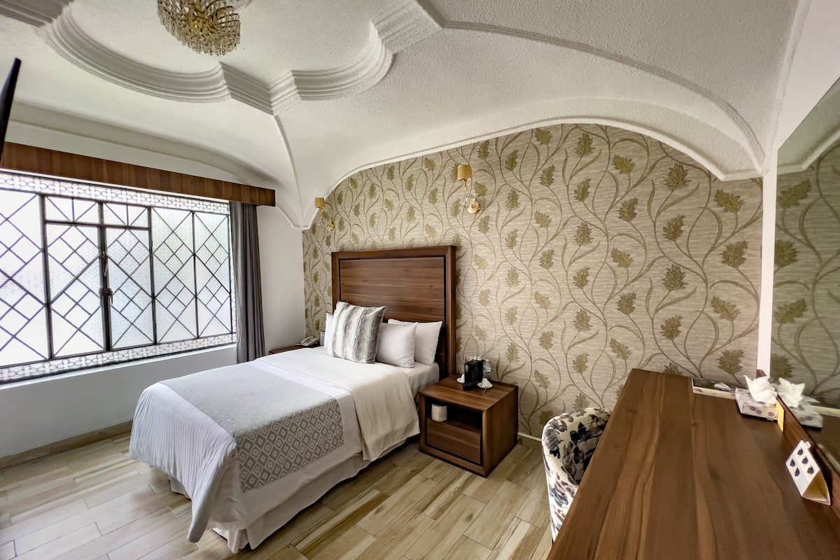 A bedroom with a large bed, a wooden nightstand, a bedside table, and a window with a view.