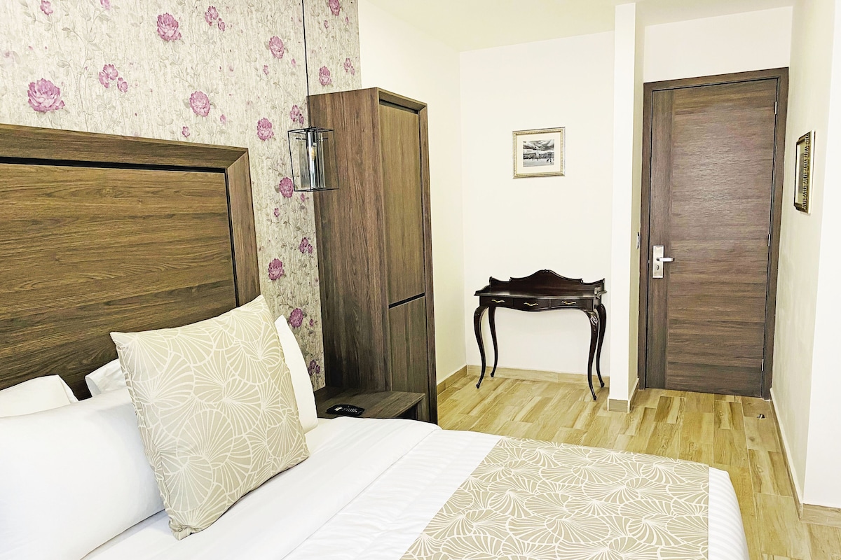 Un dormitorio con cabecera de madera, cama con ropa de cama estampada, mesita de noche, armario, una pequeña mesa y una puerta.