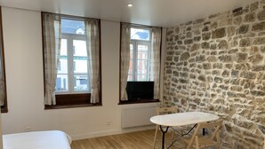 Une pièce avec un mur en pierre, un plancher en bois, un lit, une table avec une chaise et un téléviseur posé sur une étagère.