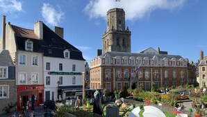 Une place pavée, bordée de bâtiments historiques, avec un beffroi et un jardin public agrémenté de sculptures.