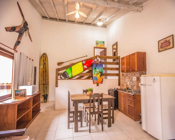 Una cocina compacta con armarios de madera, una mesa de comedor con sillas, un ventilador de techo y una tabla de surf colgada en la pared.
