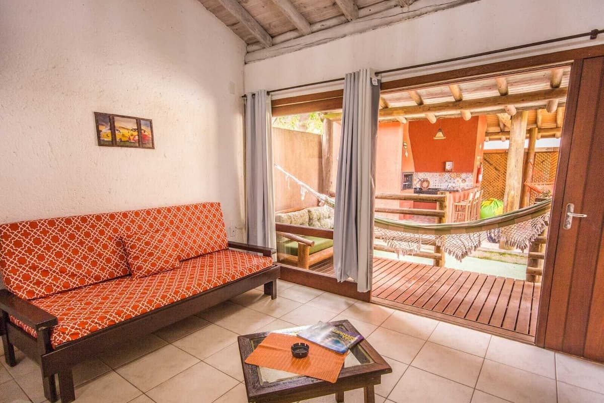 Ein Zimmer mit einer gemusterten Couch, einem kleinen Tisch mit einer Zeitschrift und Blick auf einen Außenbereich mit Holzterrasse und Hängematte.