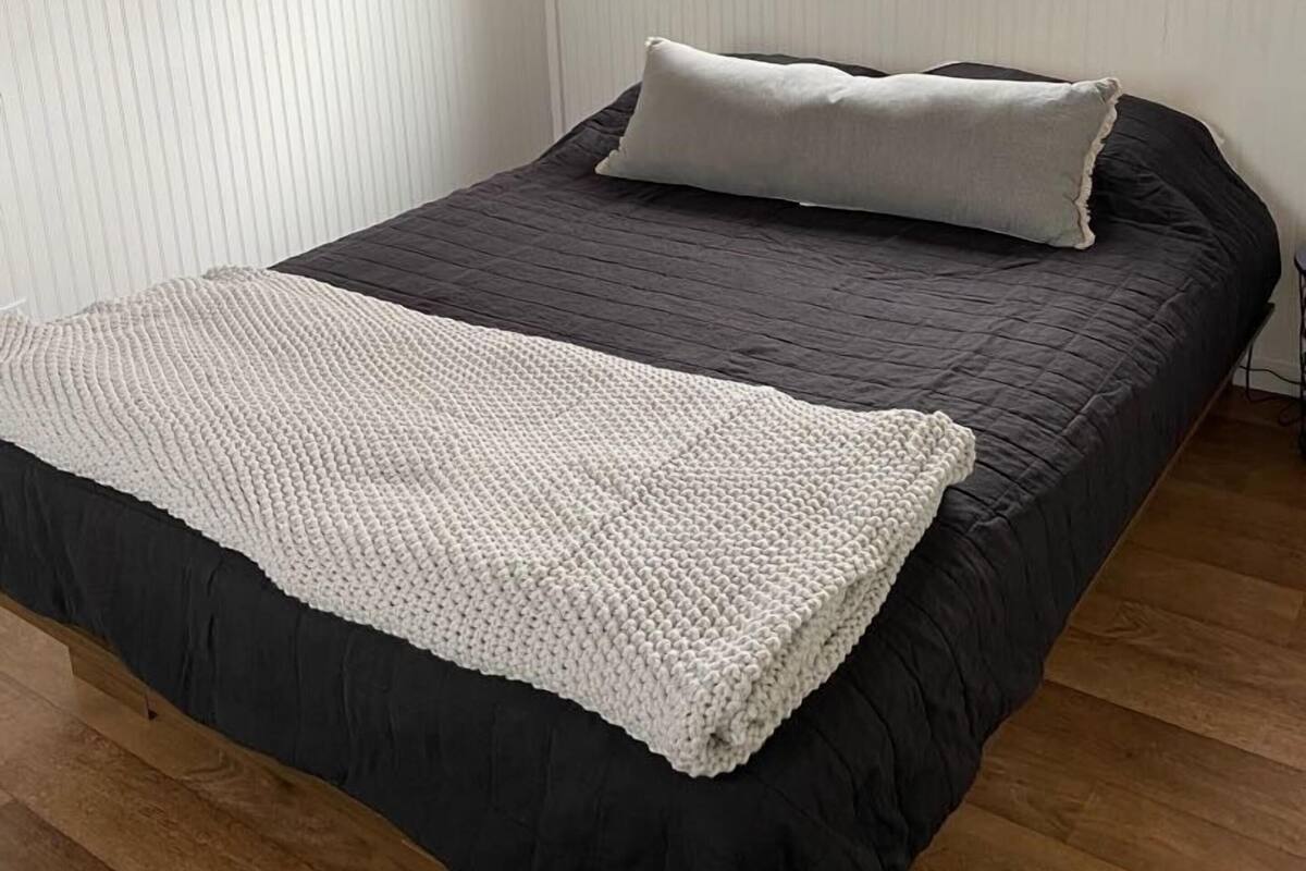 Una cama individual con un edredón oscuro y una manta blanca de punto, una ventana con persianas y suelo de madera.