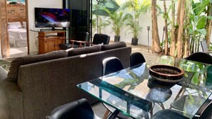 A modern living room with a glass dining table, comfortable seating, a TV, and a view of a garden with potted plants. - Villa Cocos, Modern Family Retreat, Cozy & Private just 3 Blocks from the Beach (San Francisco)
