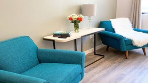 A teal armchair, a small table with books and flowers, and a lamp in a modern room with wooden flooring.
