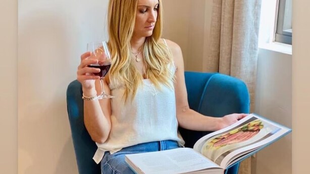 A person sitting on a blue chair, holding a glass of red wine and a book.