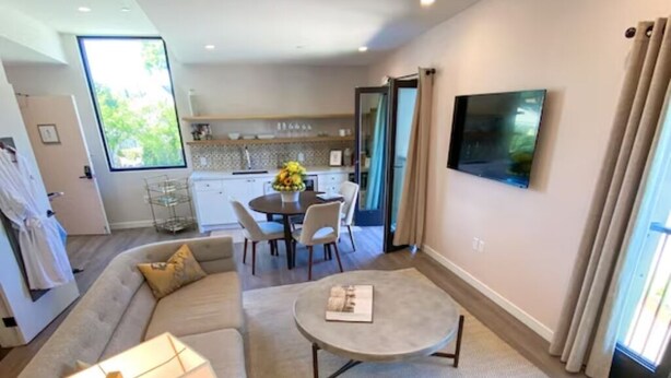 A modern living room with a sofa, a coffee table, and a TV. There is a dining area with a table and chairs in the background.