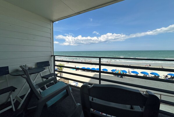A balcony with a view of the beach, featuring a table and chairs, and a railing.