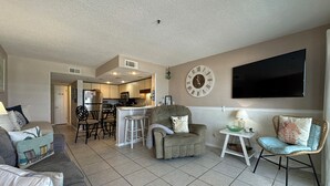 A living room with a sofa, a chair, a dining area with chairs, a kitchen with cabinets and appliances, and a wall-mounted TV.