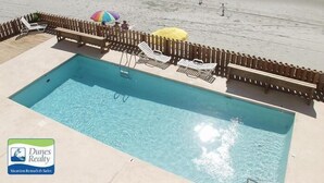 A pool area with lounge chairs and umbrellas, adjacent to a beach with people walking.