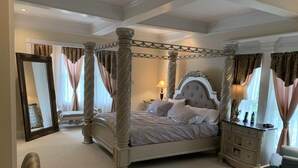 Una cama con dosel, un cabecero capitonado, una mesita de noche con lámpara y una silla en un dormitorio con un espejo grande y cortinas.