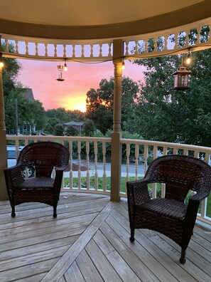 A covered porch with two wicker chairs and a railing, overlooking a road and trees.