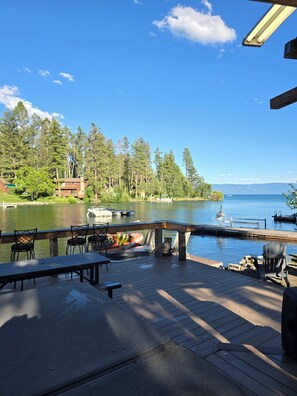 A lakeside deck with outdoor seating, a view of the lake, and a distant mountain range. - Flathead Lake home W/ private beach, dock, and hot tub (Lakeside)