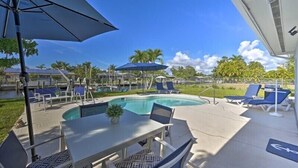 A poolside area with lounge chairs and umbrellas, a table with chairs, and a clear view of the pool. - Canelas Home, you’re Home away from Home (Cape Coral)