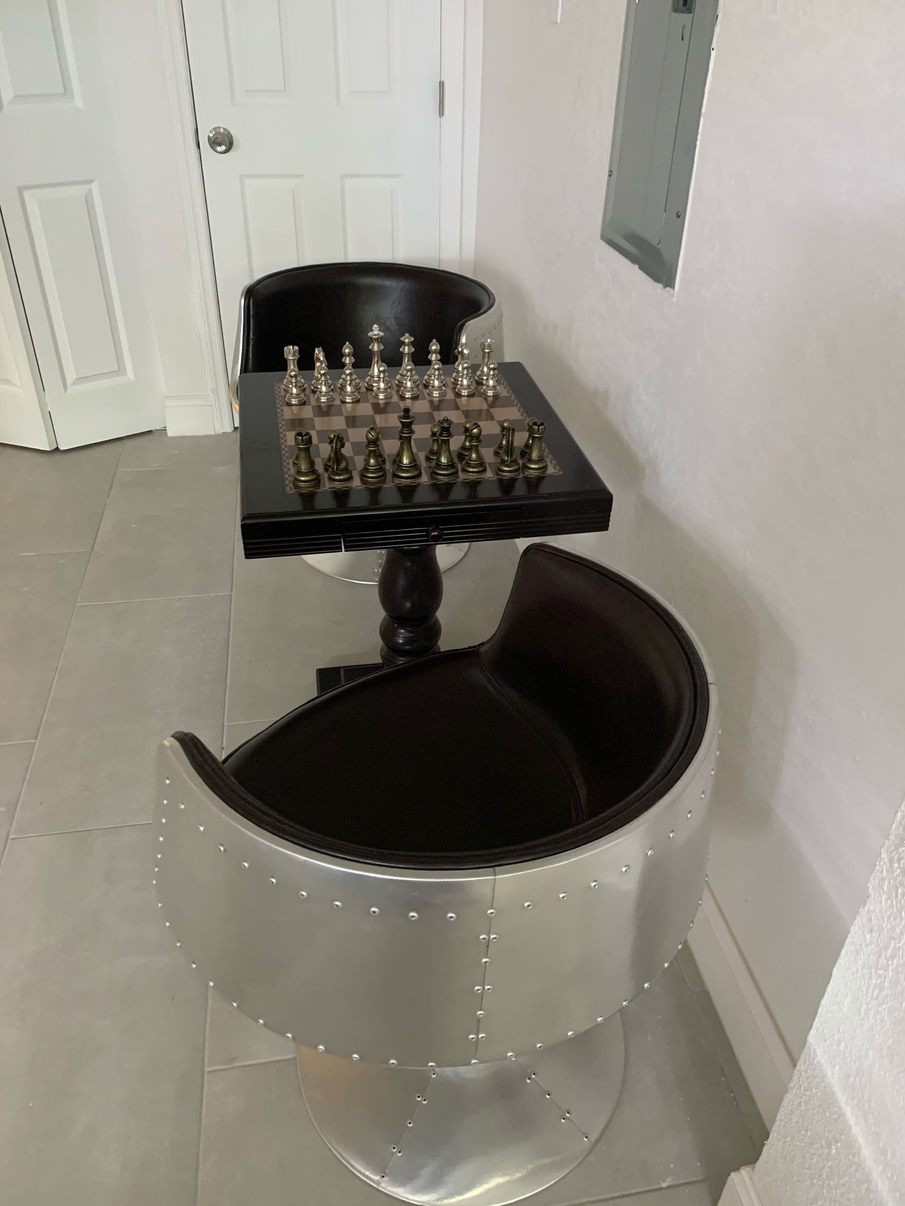 A room with a chessboard on a table, a chair with a unique design, and a white door in the background.