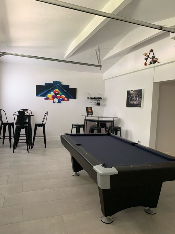 A pool table with chairs, a small table with a drink dispenser, and a wall with a billiards-themed artwork.