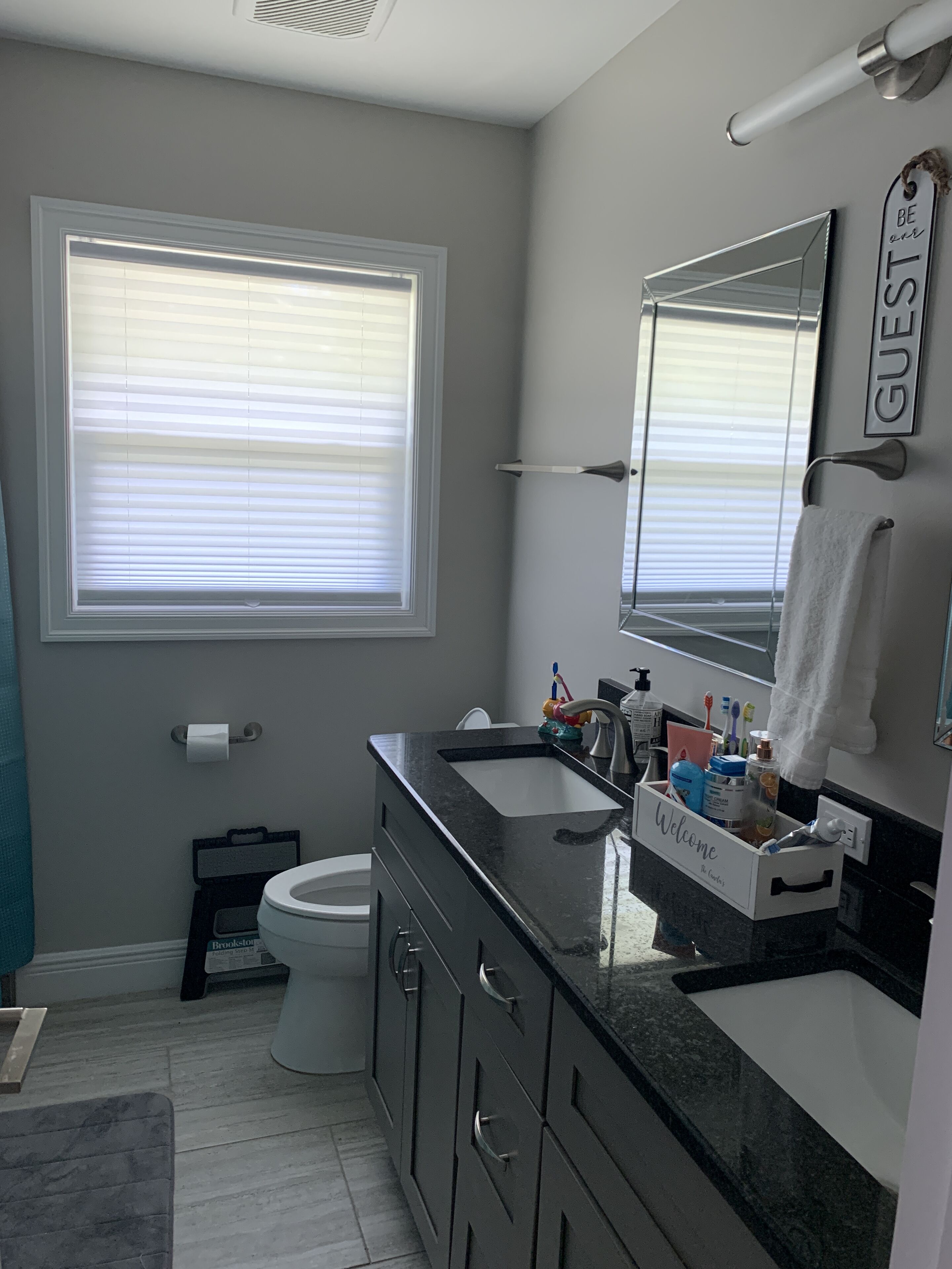 A modern bathroom with a toilet, sink, and mirror. There is a window with blinds, a towel rack, and a sign that says "GUEST".