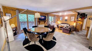 A cozy living room with a round wooden table, comfortable seating, a television, and a balcony with a view of greenery.