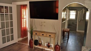 A living room with a large flat-screen TV mounted on the wall, a wooden console table with decorative vases, and a staircase leading to another level. - The Bungalow: inviting retreat with a spacious backyard sets for BBQs.  (Grand Forks)