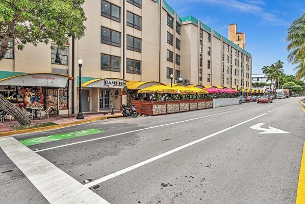 A street view with a row of outdoor seating areas with umbrellas, a building with a sign reading "HAMILTON," and a clear road with directional arrows.