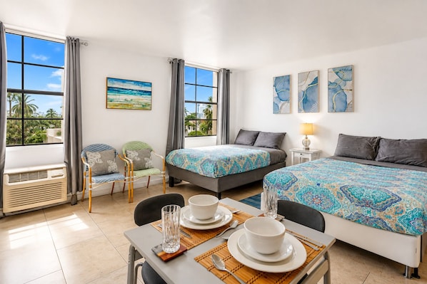 A hotel room with two beds, a dining table with plates and cutlery, a chair, a window with curtains, and artwork on the walls.