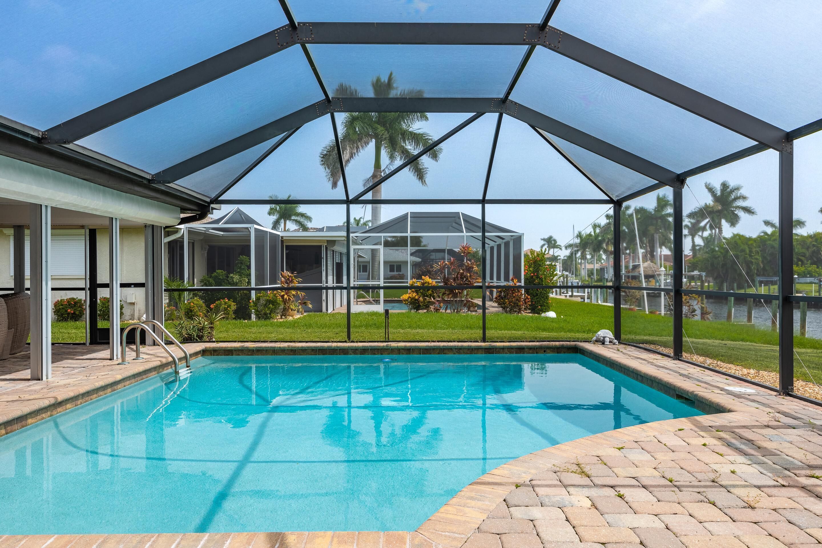 Une piscine couverte avec une structure en verre, un espace patio et une vue sur une maison et des palmiers.