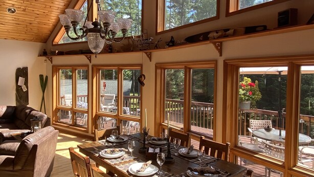 Uma ampla área de jantar com teto de madeira alto e grandes janelas, uma mesa de jantar de madeira para quatro pessoas, e vista para um deck com móveis de exterior.