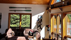 Uma sala de estar ampla com teto de madeira rústica, um lustre feito de chifres e vista para árvores através de grandes janelas.