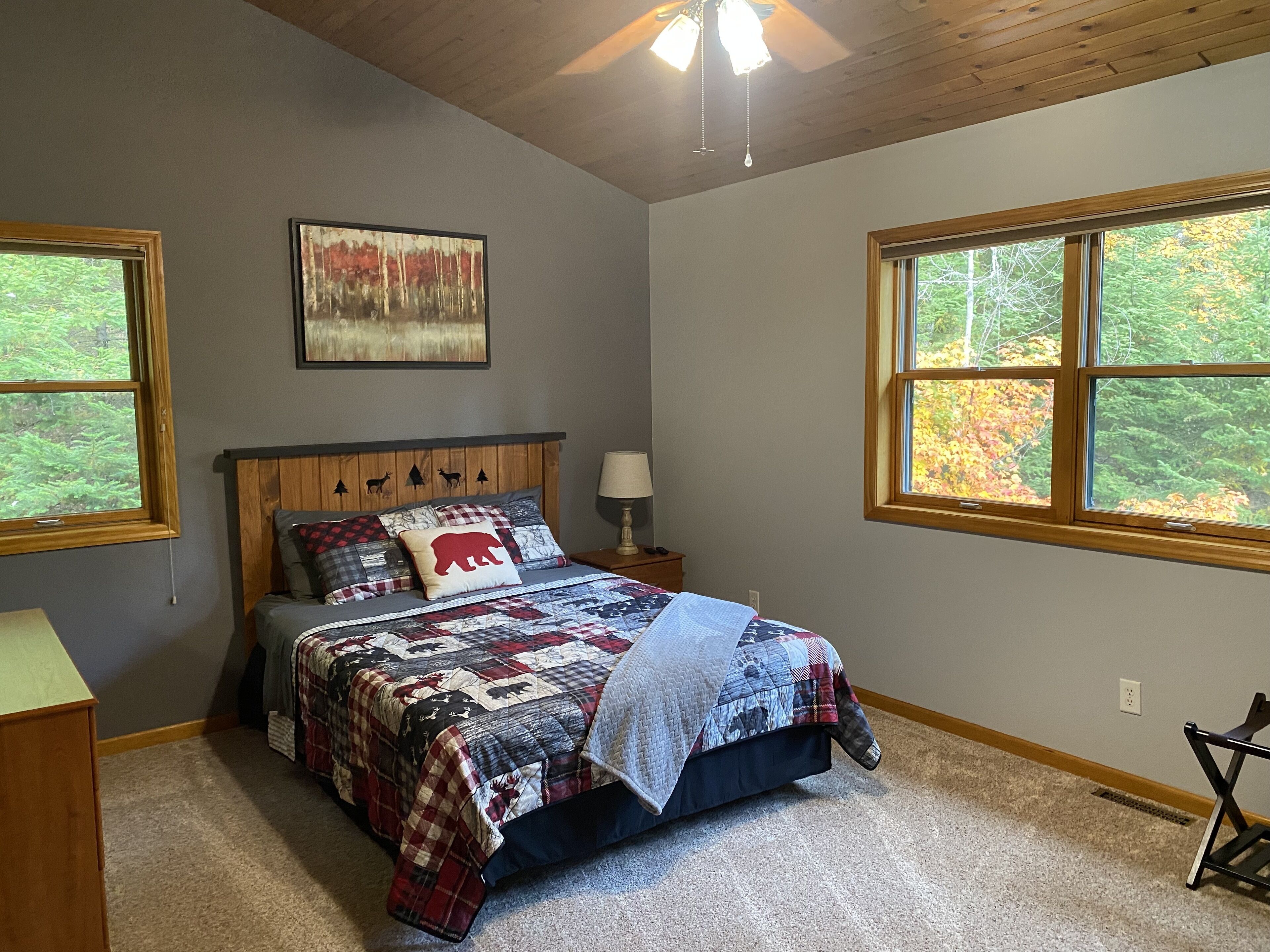 A bedroom with a wooden headboard, a bed with a quilt, two windows with a view of trees, and a ceiling fan.