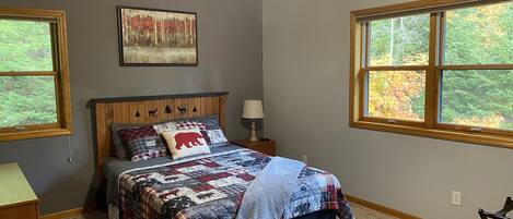 A bedroom with a wooden headboard, a bed with a quilt, two windows with a view of trees, and a ceiling fan.
