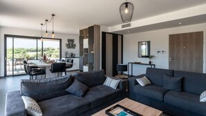 Ein modernes Wohnzimmer mit dunklen Sofas, einem Couchtisch und Pendelleuchten. Durch eine Glastür ist ein Essbereich mit großem Tisch und Stühlen zu sehen.