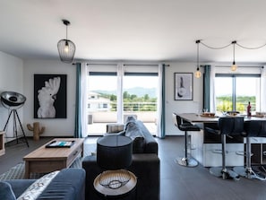 Un salon moderne avec un canapé, une table basse et un coin repas avec des chaises. De grandes fenêtres offrent une vue sur l’extérieur.