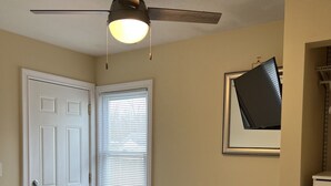 Une chambre à coucher comprenant un lit, un ventilateur de plafond, une fenêtre avec des stores, un téléviseur fixé au mur et une commode.