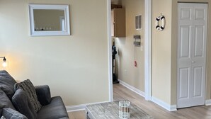 Un salon avec un canapé gris, une table basse en bois, un ventilateur de plafond, un miroir et une porte sur laquelle est accroché un gilet de sauvetage.