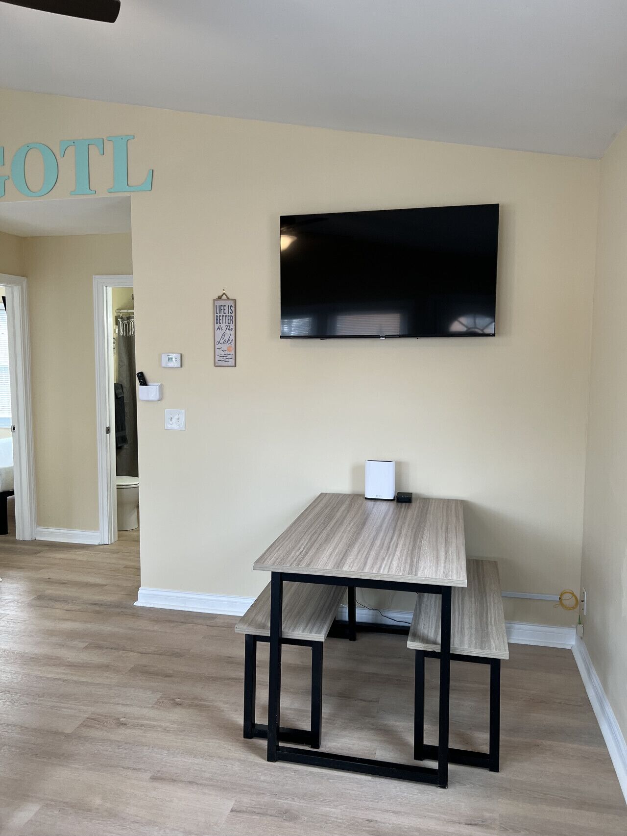A living room with a wooden table and bench, a flat-screen TV mounted on the wall, and a door leading to another room.