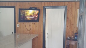 Habitació petita amb paret de fusta, ventilador de sostre, televisió penjada a la pared, una porta i una taula petita amb una cadira.