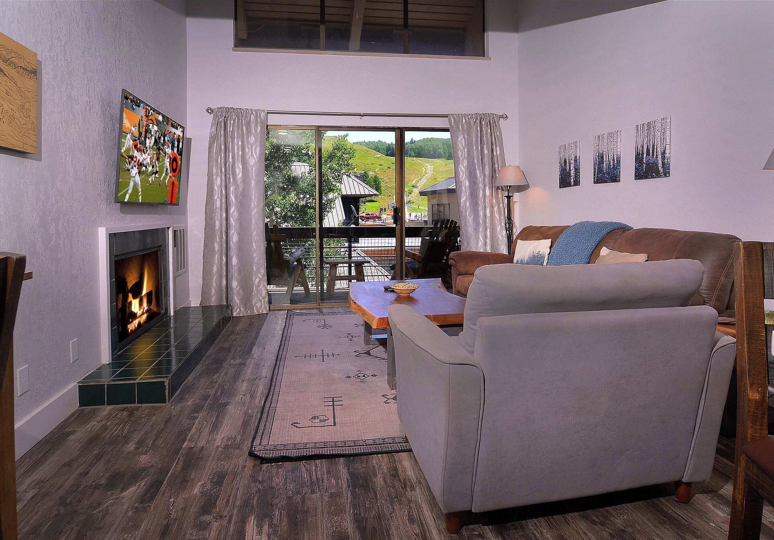 A living room with a fireplace, a sofa, a coffee table, and a view of a green landscape through large windows.