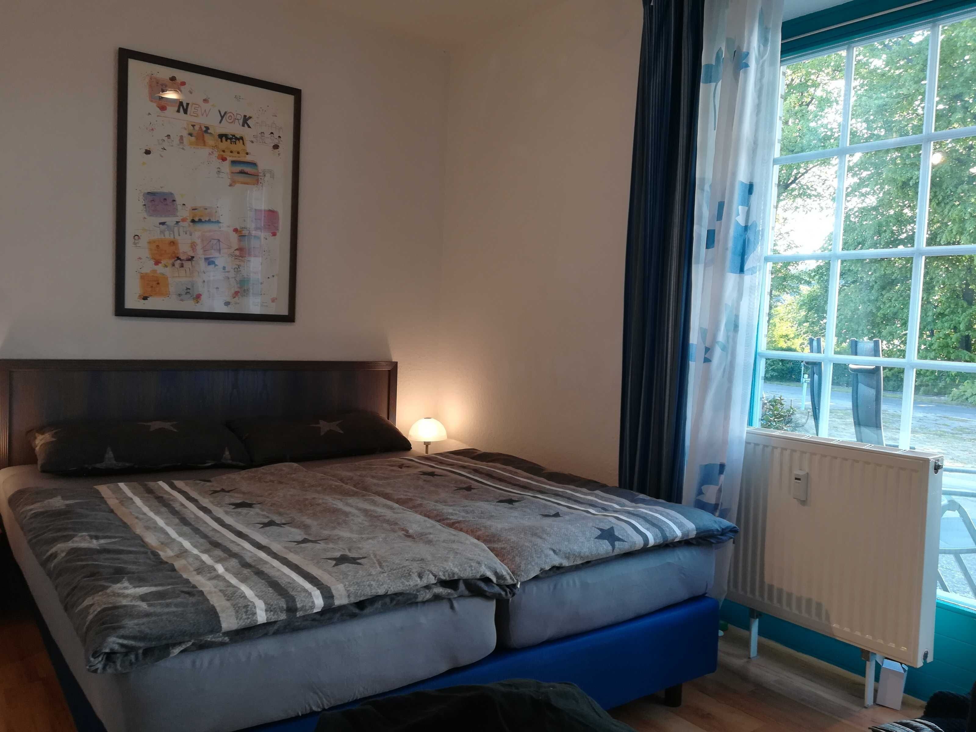 A bedroom with a bed, a nightstand, a lamp, a window with curtains, and a framed picture on the wall.