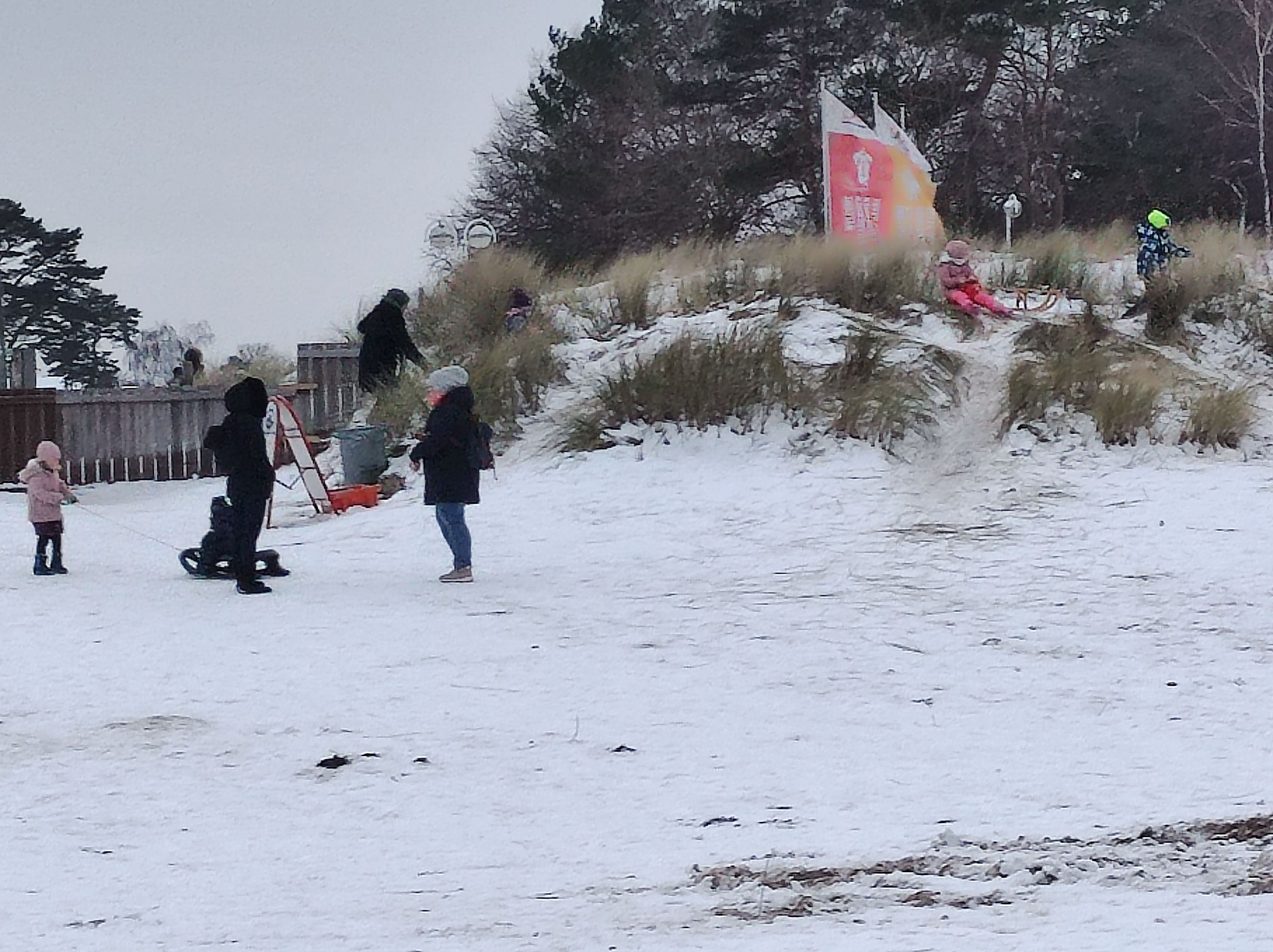 A snowy outdoor area with people, including children, and a flag on a hill.