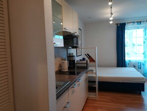 Et lille værelse med tekøkken, seng og et vindue med gardiner.