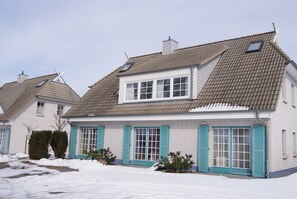 Una casa de dos pisos con techo a dos aguas, ventanas dormerunas y un porche sobre suelo cubierto de nieve.