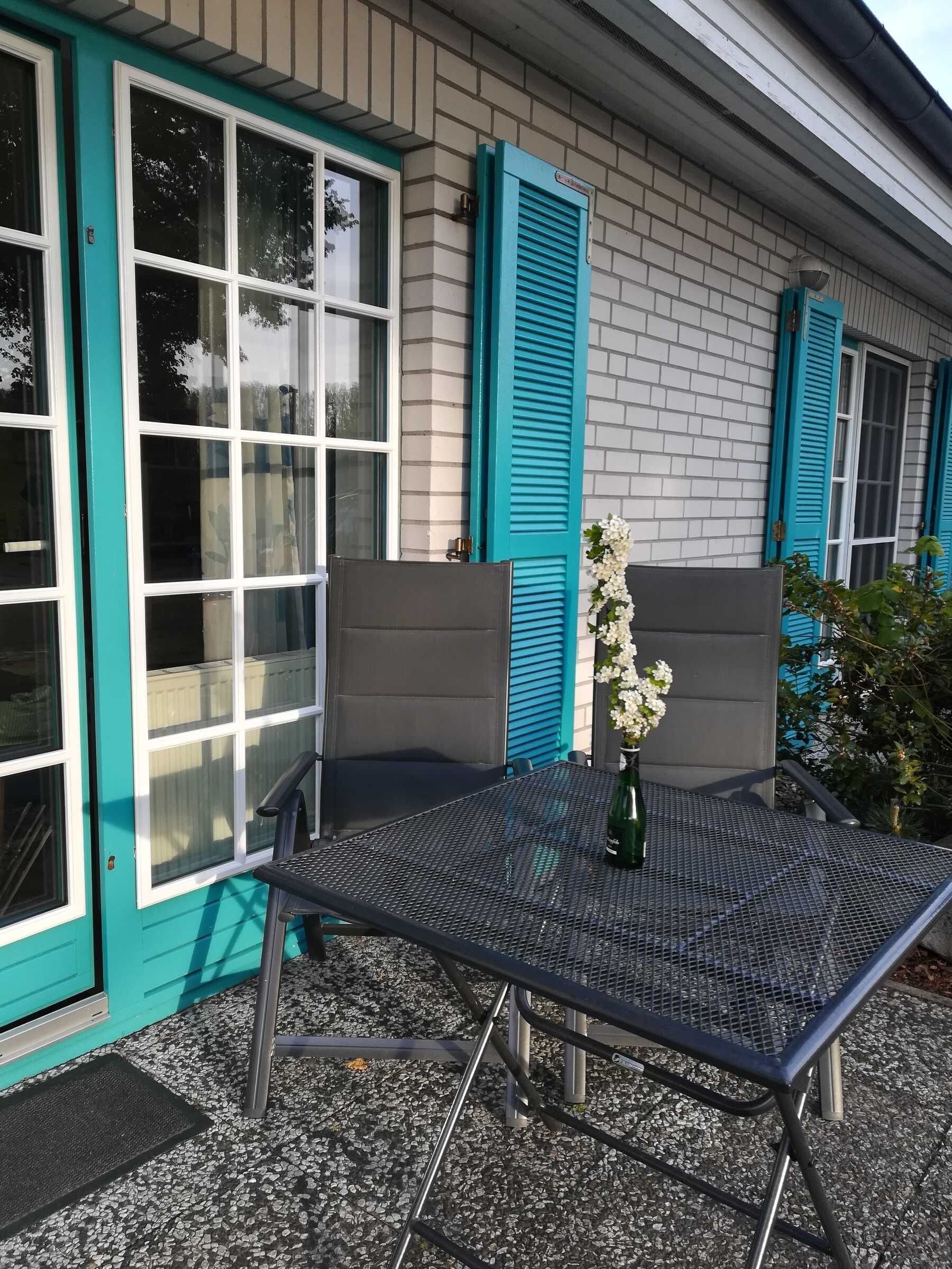A patio with a metal table and chairs, a potted plant, and a building with teal shutters and windows.