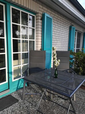 En terrasse med et metalbord og stole, en potteplante og en bygning med turkise skodder og vinduer.