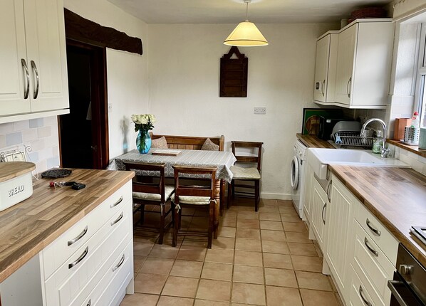 A compact kitchen with white cabinets, a wooden countertop, a dining table with chairs, a washing machine, and a hanging light fixture. - Pretty cottage, perfect readers' retreat in centre of Hay, parking & fast wifi (Hay-on-Wye)
